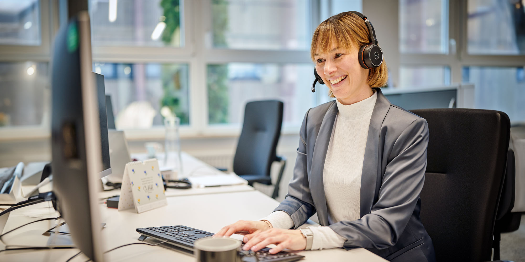 A smiling woman working at an office desk wearing a headset. 